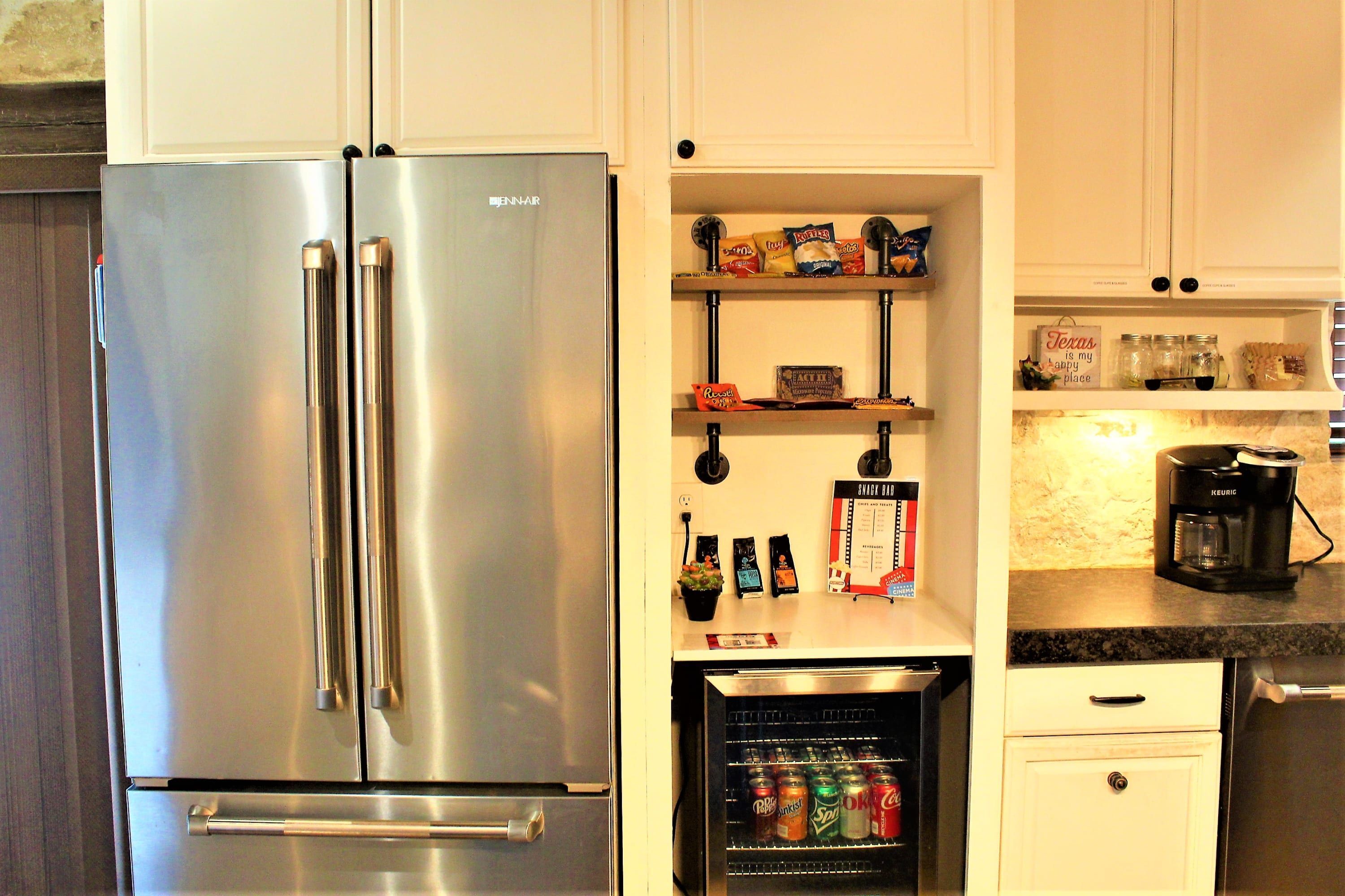 A kitchen with a stainless steel refrigerator, a beverage cooler, shelves with snacks, and a coffee maker.