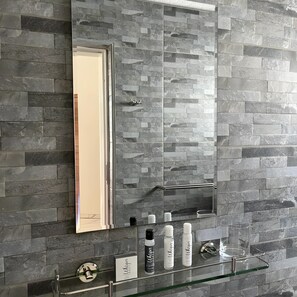 A bathroom with a stone wall, a glass shelf with toiletries, and a mirror. - The Baroness (Bulawayo)