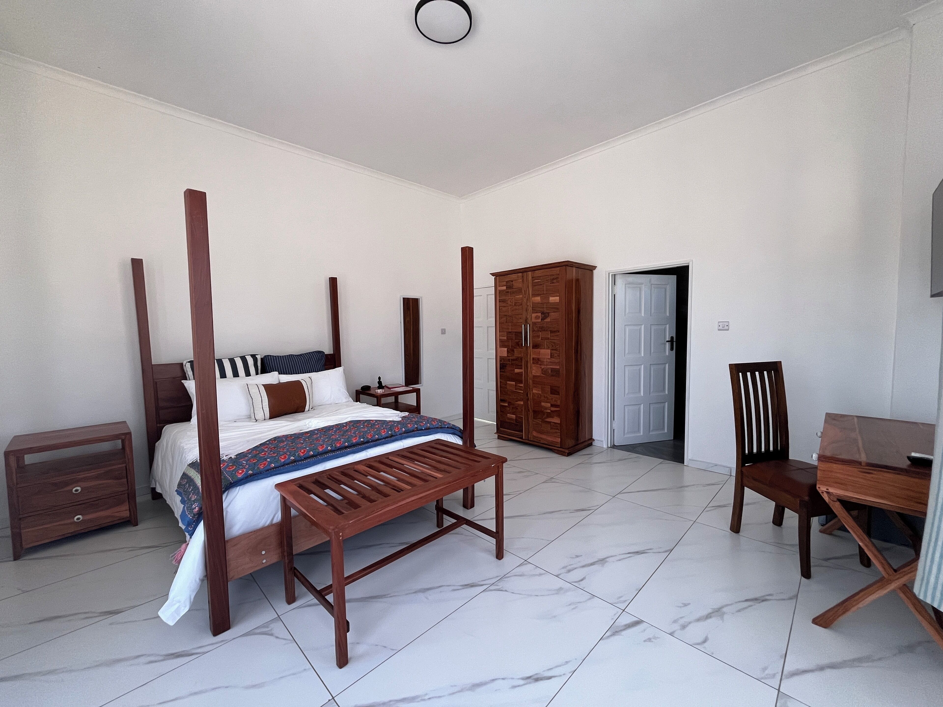 A bedroom with a four-poster bed, a wooden wardrobe, a desk with a chair, and a small bedside table.