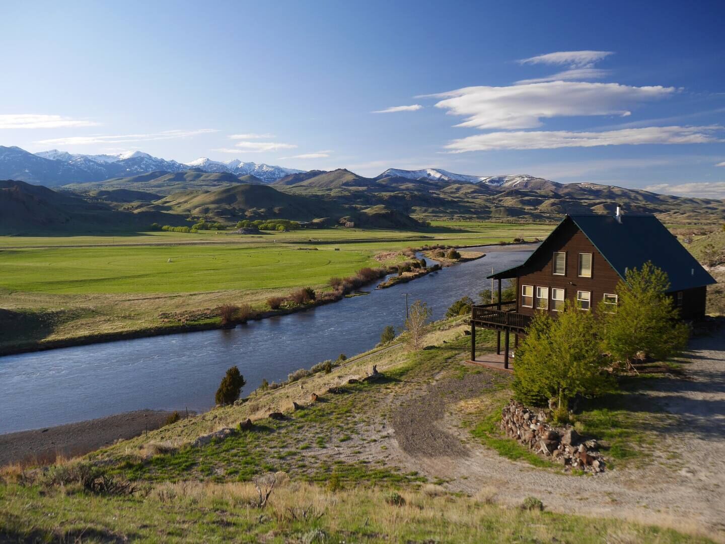 A house with a dark roof and multiple windows, situated on a hillside with a river in the foreground and mountains in the background.