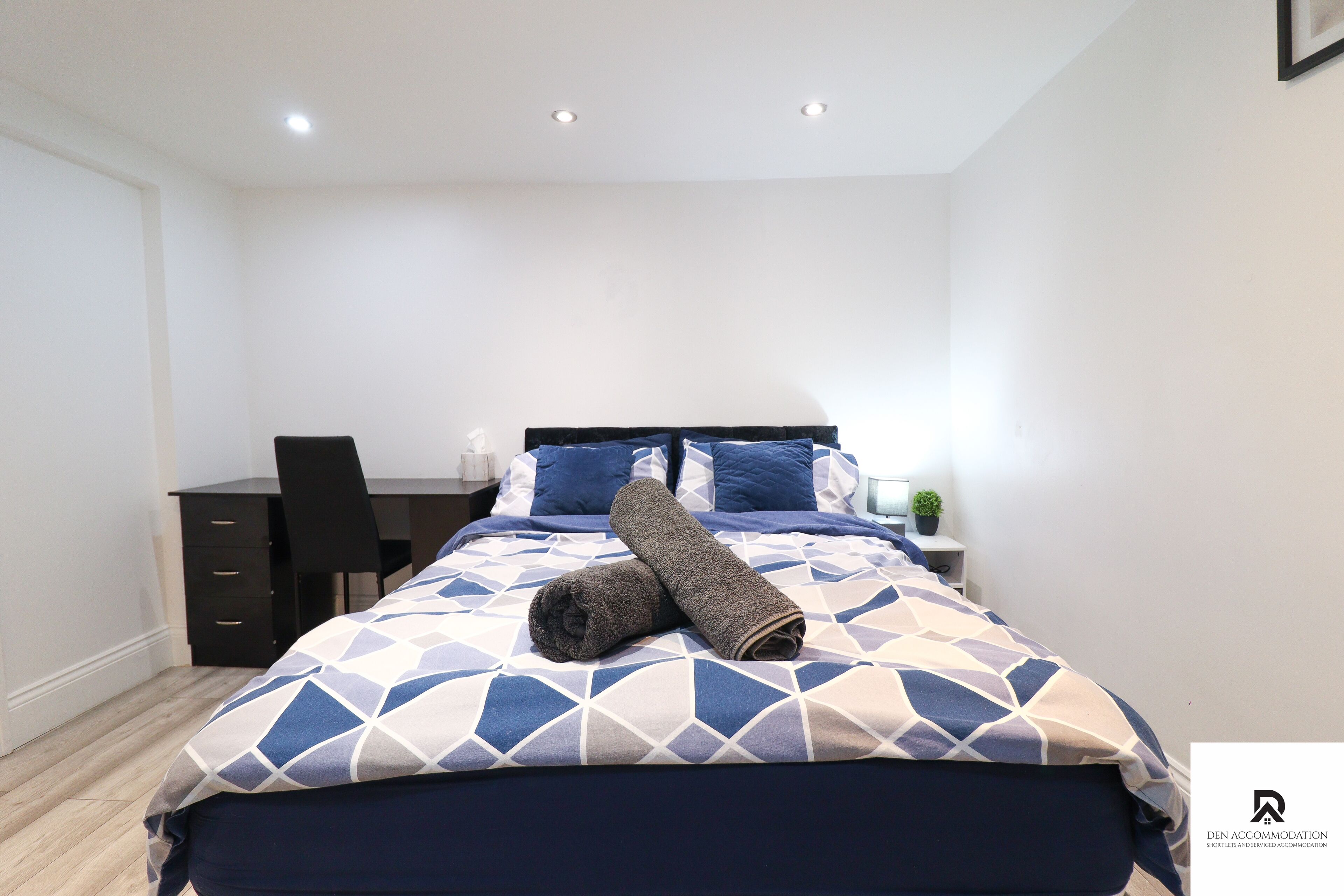 A neatly made bed with a geometric-patterned duvet, a rolled-up towel, and matching pillows. There is a small bedside table with a lamp and a potted plant. A dark-colored desk chair is positioned next to the bed.