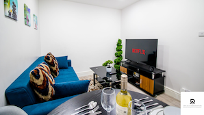 A modern living room with a blue sofa, a dining table set with cutlery and a bottle of wine, a flat-screen TV, and a small potted plant. - 2-bed Near the O2 Arena & Excel & Free Parking (London)