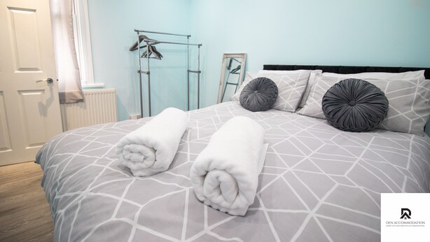A neatly made bed with rolled towels, a radiator, a drying rack, and a mirror on the wall. - 2-bed Near the O2 Arena & Excel & Free Parking (London)