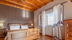 A bedroom with a wooden bed, a nightstand, a lamp, a window with curtains, and a view of the sea.
