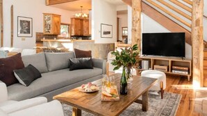 Amplia sala de estar con una escalera alta de madera, un sofá gris, una mesa de centro de madera con aperitivos y un televisor.