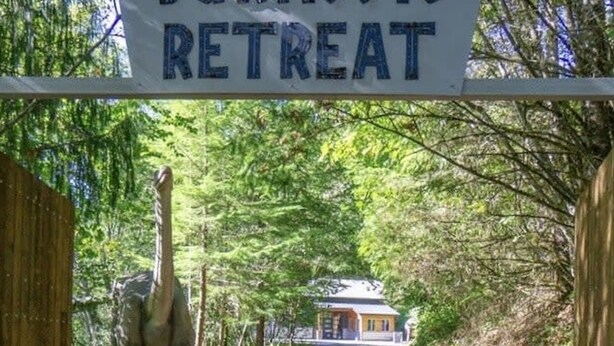 A sign reading "Jurassic Retreat" at the entrance of a path leading to a house surrounded by trees.