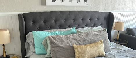 A neatly made bed with a grey headboard, multiple pillows, and a framed surfboard art piece above it.