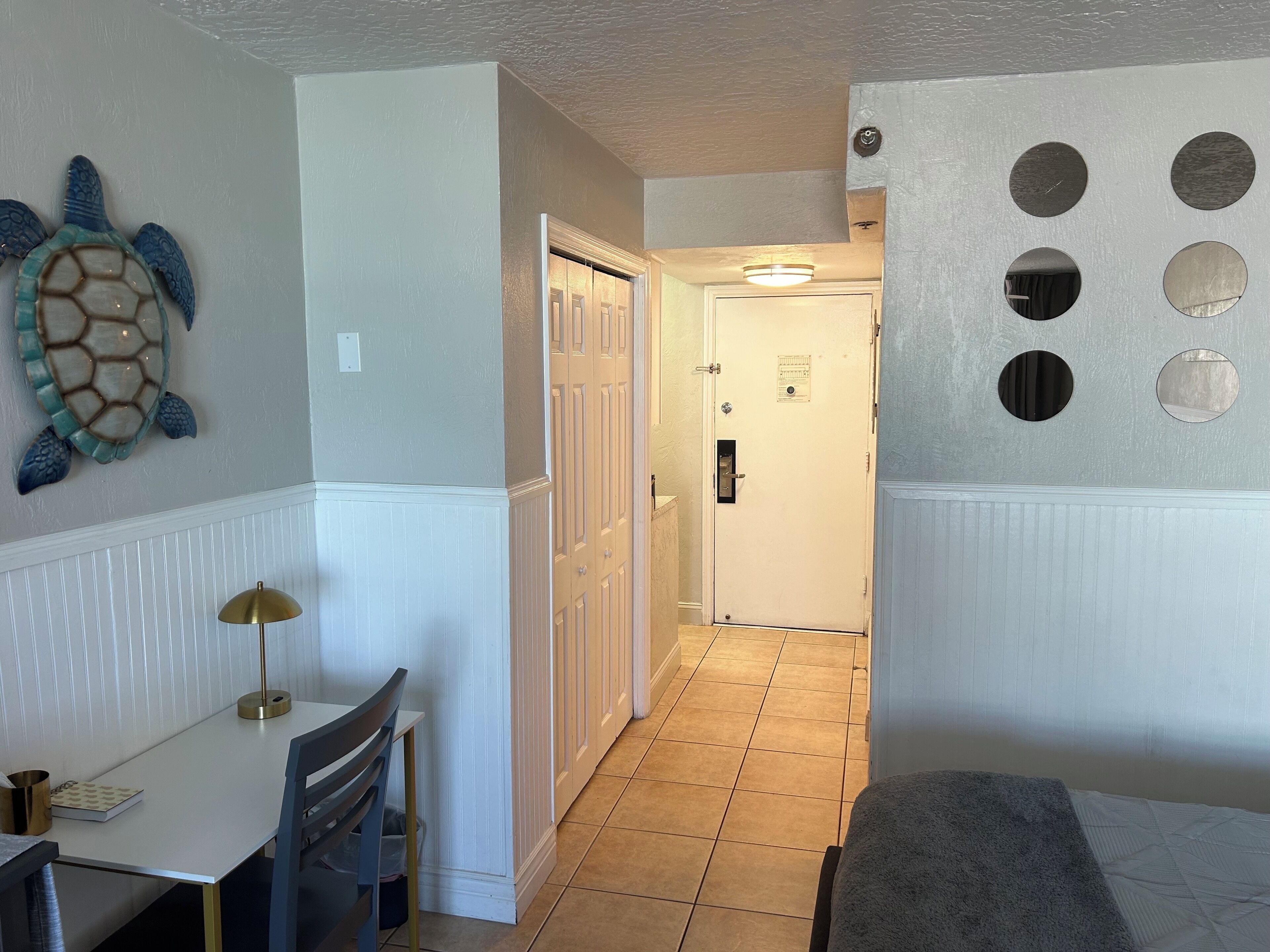 A hallway with a white door, a wall-mounted turtle decoration, a table with a lamp, and a bed with a gray blanket.