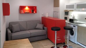 A compact living space with a grey sofa, a red accent wall, a kitchen area with a washing machine, and a small bar stool.