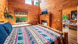 A cozy log cabin bedroom with a bed, a fireplace, a television, and a balcony with a view of the outdoors. - Hot tub|Fireplace|FastWiFi|Convenient|Pet Friendly (Pigeon Forge)