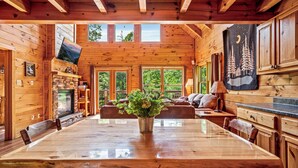 A wooden cabin interior with a dining table, chairs, a fireplace, a TV, and a large window overlooking greenery. - Hot tub|Fireplace|FastWiFi|Convenient|Pet Friendly (Pigeon Forge)