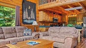A cozy cabin interior with two sofas, a wooden coffee table, and a kitchen area with wooden cabinets and a large window. - Hot tub|Fireplace|FastWiFi|Convenient|Pet Friendly (Pigeon Forge)