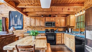 A wooden cabin interior with a dining table, chairs, a kitchen with wooden cabinets, a microwave, stove, and refrigerator. - Hot tub|Fireplace|FastWiFi|Convenient|Pet Friendly (Pigeon Forge)