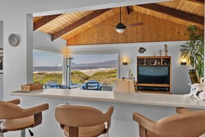 A modern kitchen with a large window overlooking a beach, a bar area with chairs, and a television. - Lands End - Ocean Front w/Beach Path & Spa (Gold Beach)