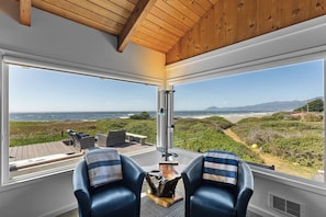 Ein Zimmer mit großem Fenster, das auf einen Strand und das Meer blickt, ausgestattet mit zwei Ledersesseln und einem kleinen Tisch.