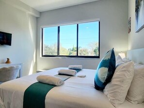 A hotel room with a large window, a bed with white linens, a green throw, and a blue pillow.