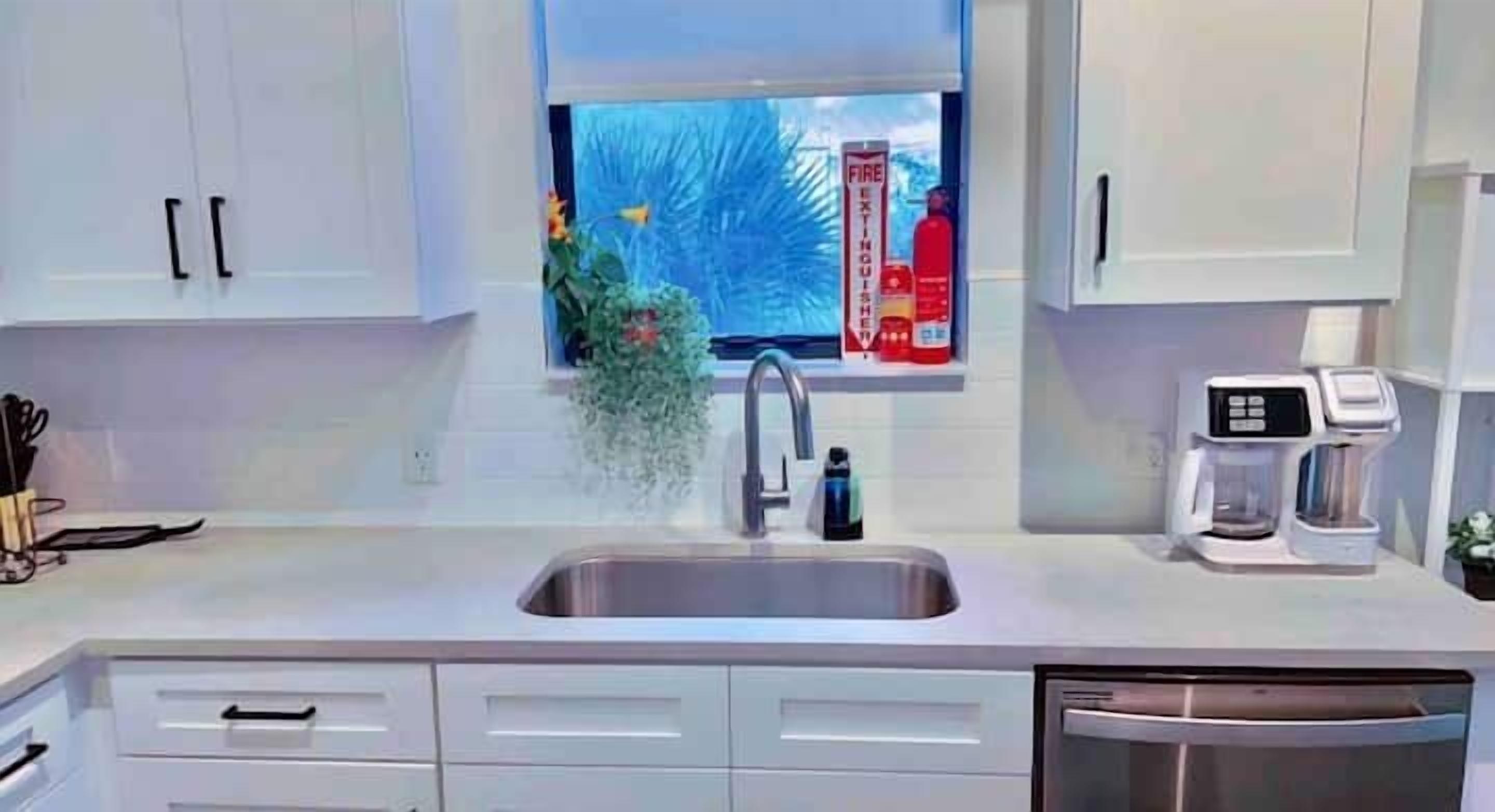 Een moderne keuken met witte kastjes, een roestvrijstalen spoelbak, een koffiezetapparaat en een raam met uitzicht op palmbomen.