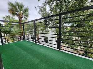 A balcony with artificial green turf, metal railings, and a view of surrounding trees and buildings.