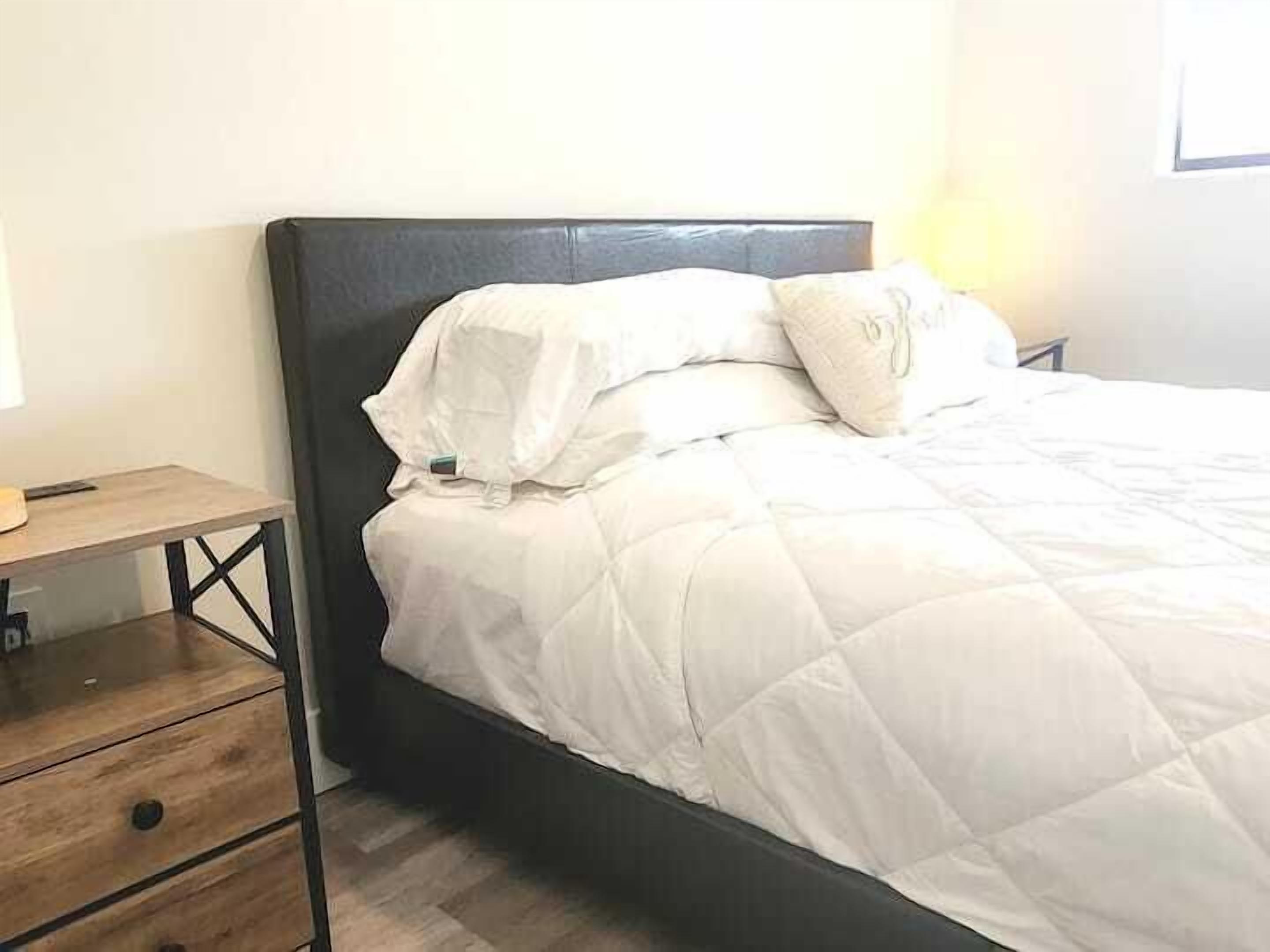 Uma cama com colcha branca, uma mesa de cabeceira de madeira com gavetas e uma cabeceira escura.