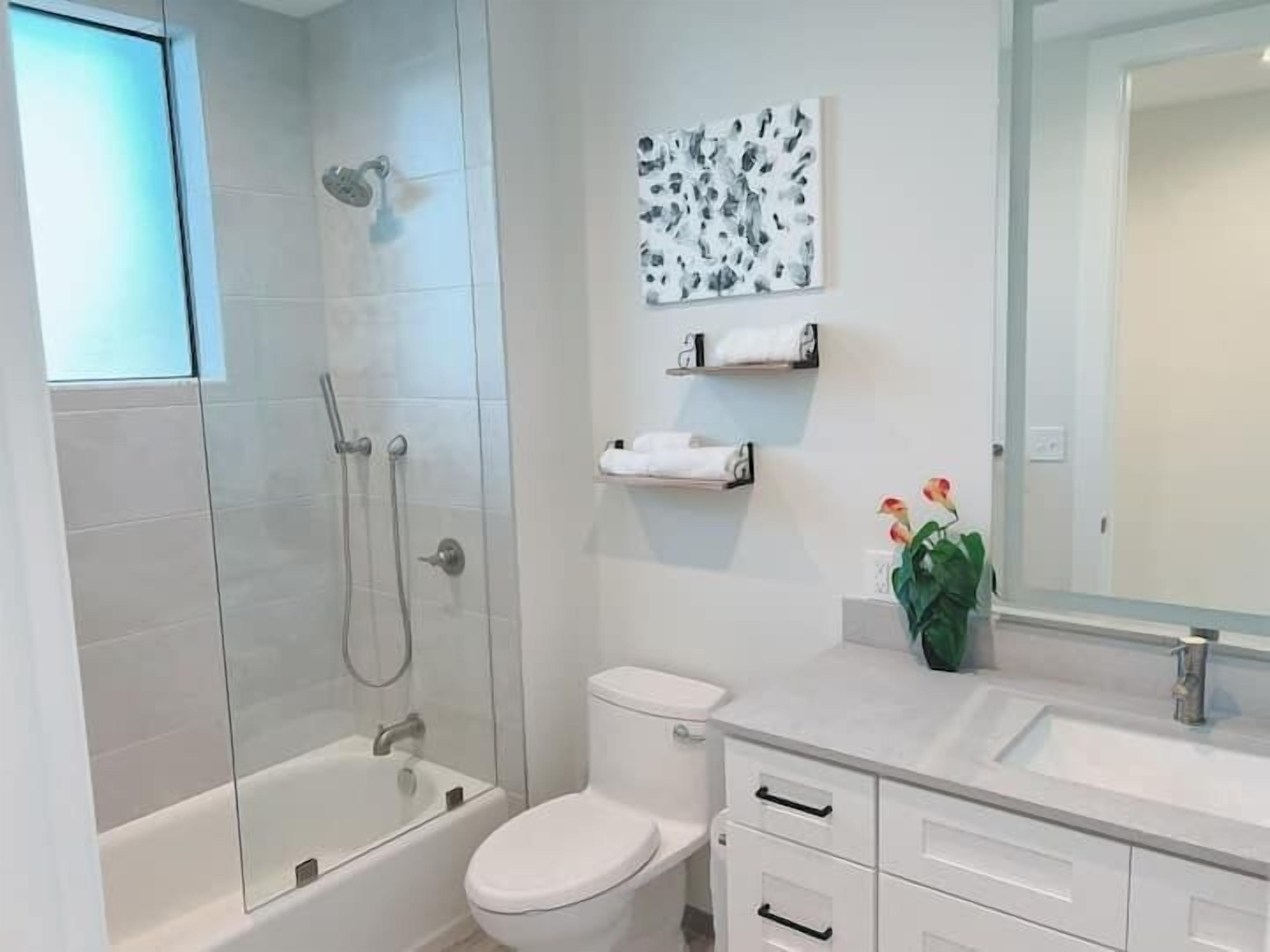 Un baño moderno con ducha de vidrio, inodoro blanco, lavamanos con espejo y una pequeña estantería con toallas.