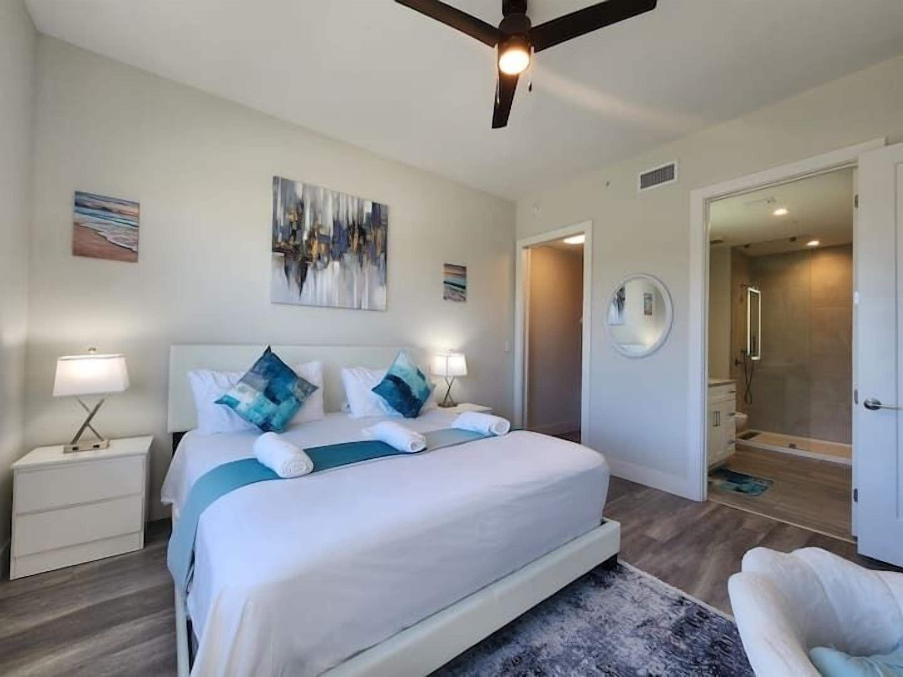 A modern hotel room with a large bed, two bedside tables with lamps, a ceiling fan, a shower area, and a round mirror on the wall.