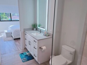 A modern bathroom with a white sink, mirror, and toilet. There is a small plant on the sink counter and a blue rug on the floor.