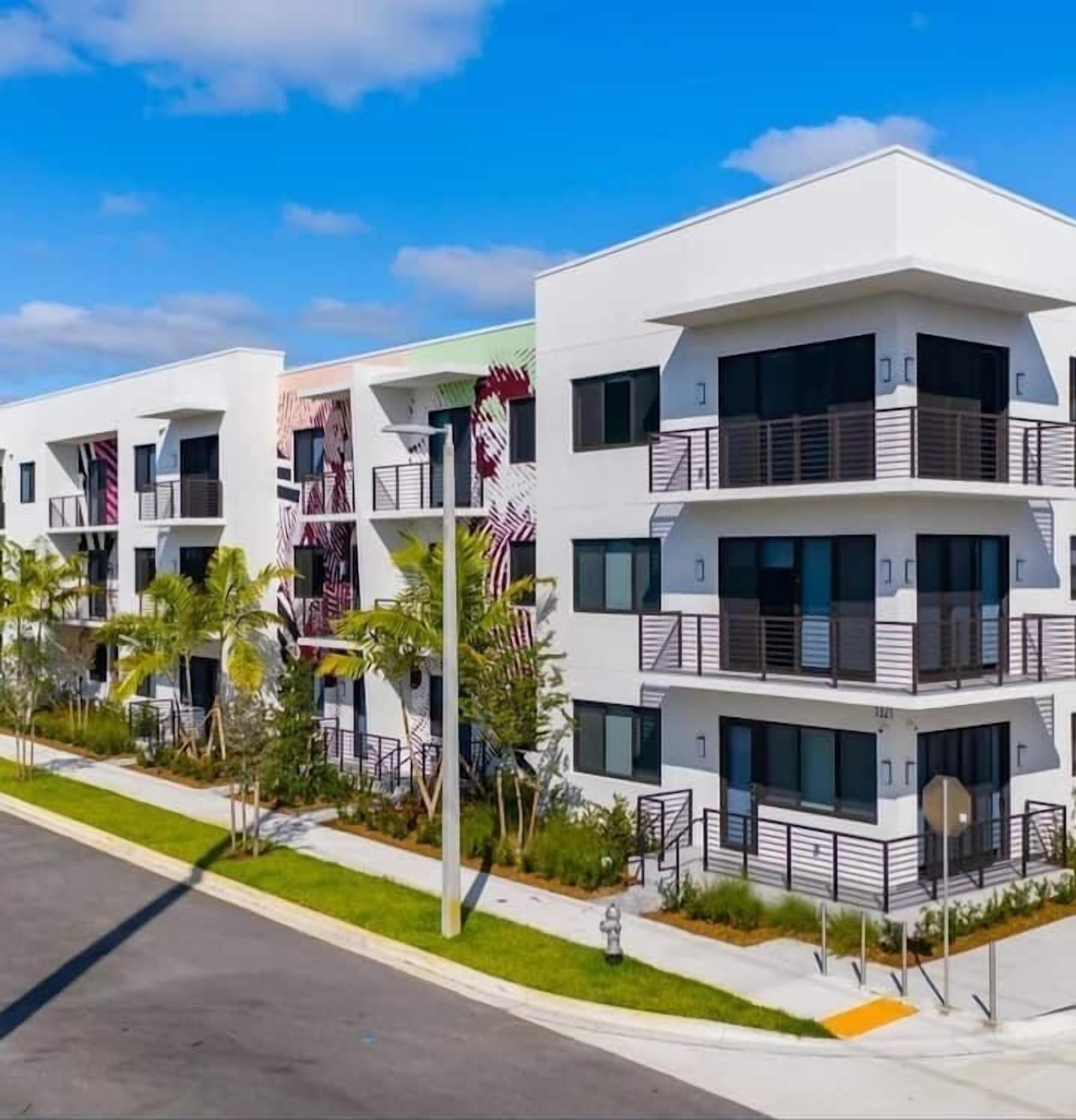 A modern multi-story residential building with balconies, a flat roof, and a landscaped front yard.