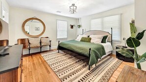 A bedroom with a bed, a desk, a chair, a mirror, and a large plant.