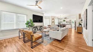 A spacious living area with a wooden floor, a ceiling fan, a flat-screen TV mounted on the wall, and a kitchen in the background.