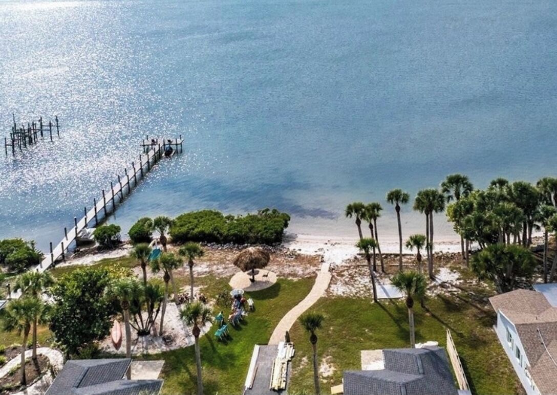 An aerial view of a beach resort with a pier extending into the water, surrounded by palm trees and a pathway leading to the beach.