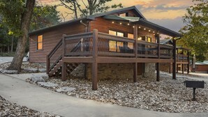 A rustic cabin with a wooden exterior, a stone foundation, and a covered deck with string lights. - Peaceful Family Lake House near SDC Walk to Lake (Branson)