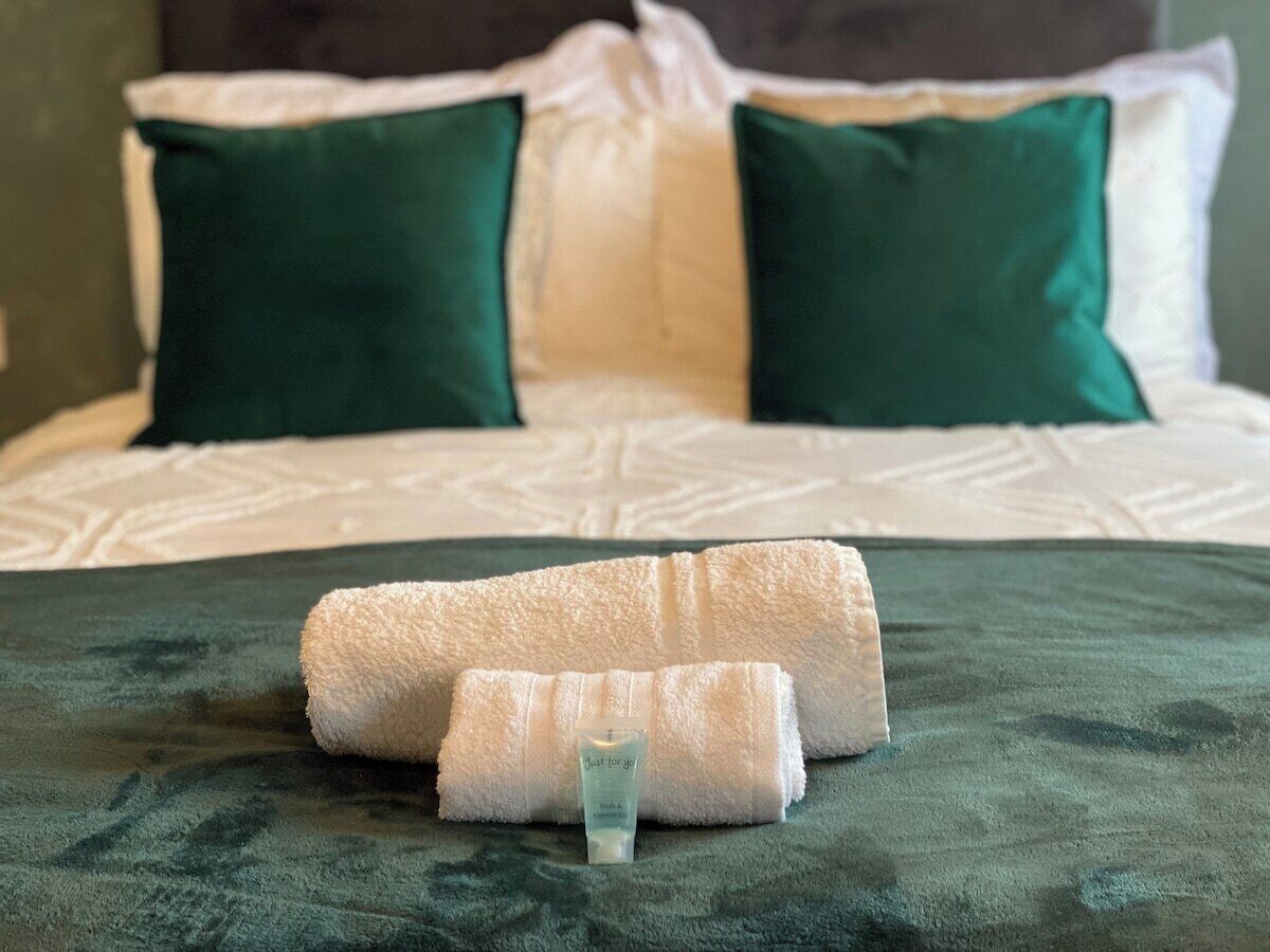 Un lit bien fait, recouvert d’une couverture verte, avec deux oreillers verts et une serviette blanche enroulée.