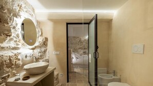 A bathroom with a stone wall, a round mirror, a sink, a bidet, and a toilet. - SUITE LE GROTTE - suite with heated pool and whirlpool (Ostuni)