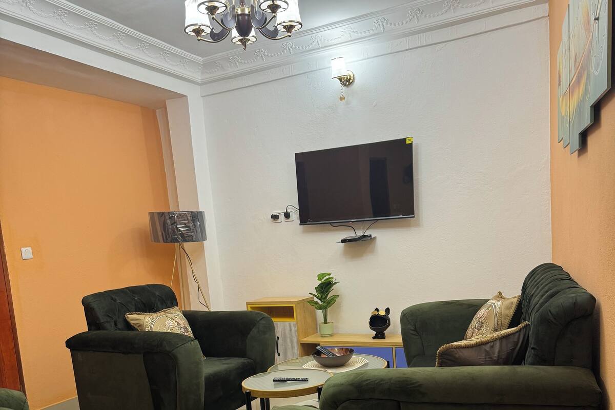 Sala de estar con sofás verdes, un televisor montado en la pared, una araña y una pequeña mesa con un cuenco y un control remoto.
