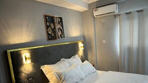 A neatly made bed with white linens, a wooden nightstand, a wall-mounted air conditioner, and two framed artworks above the bed. - Lekker Estate (Yaoundé)