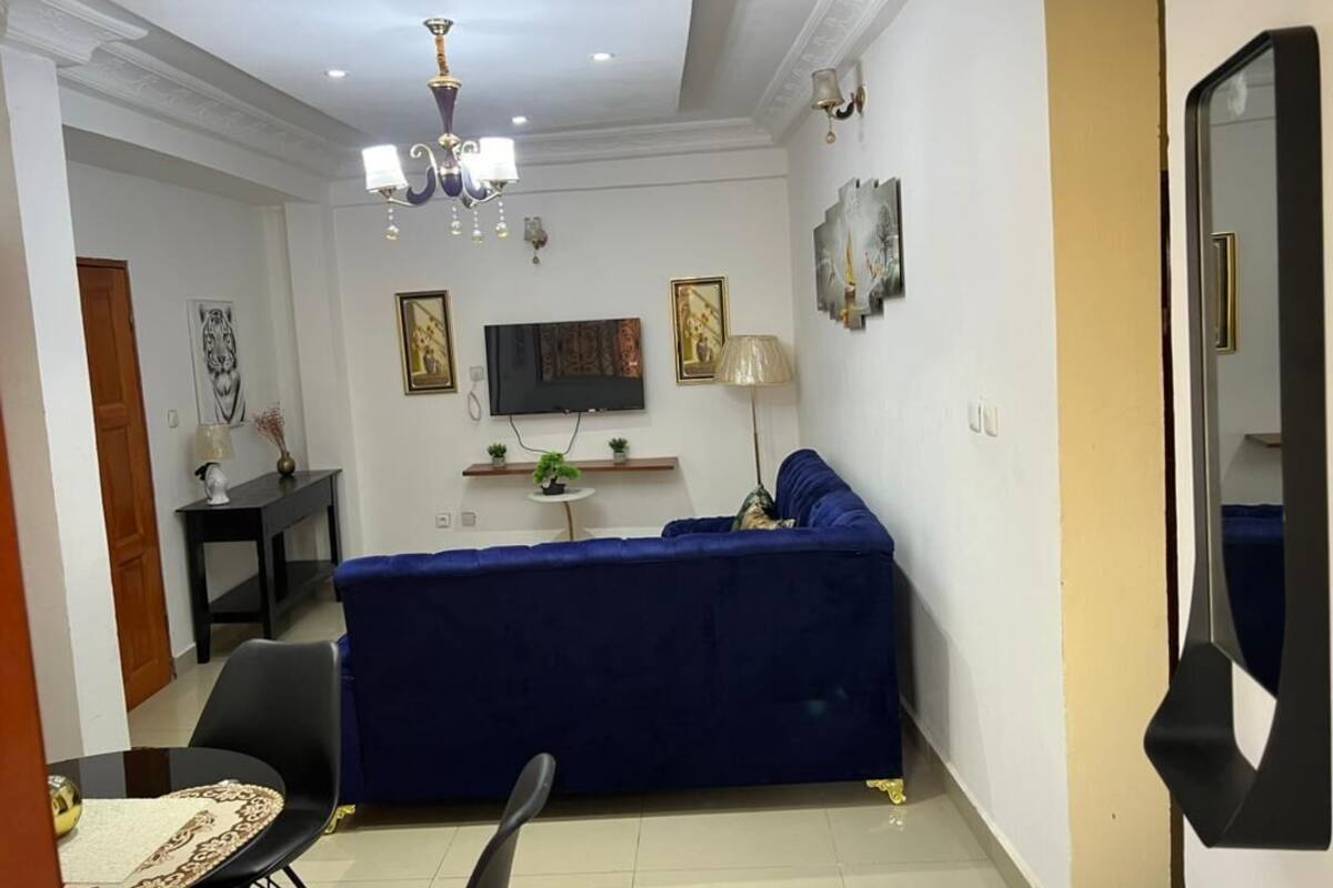 Una sala de estar con un sofá azul, un televisor, una araña y una zona de comedor con una mesa y sillas.