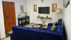 A living room with a blue sofa, a TV mounted on the wall, a small table with a plant, and a wooden door. - Lekker Estate (Yaoundé)