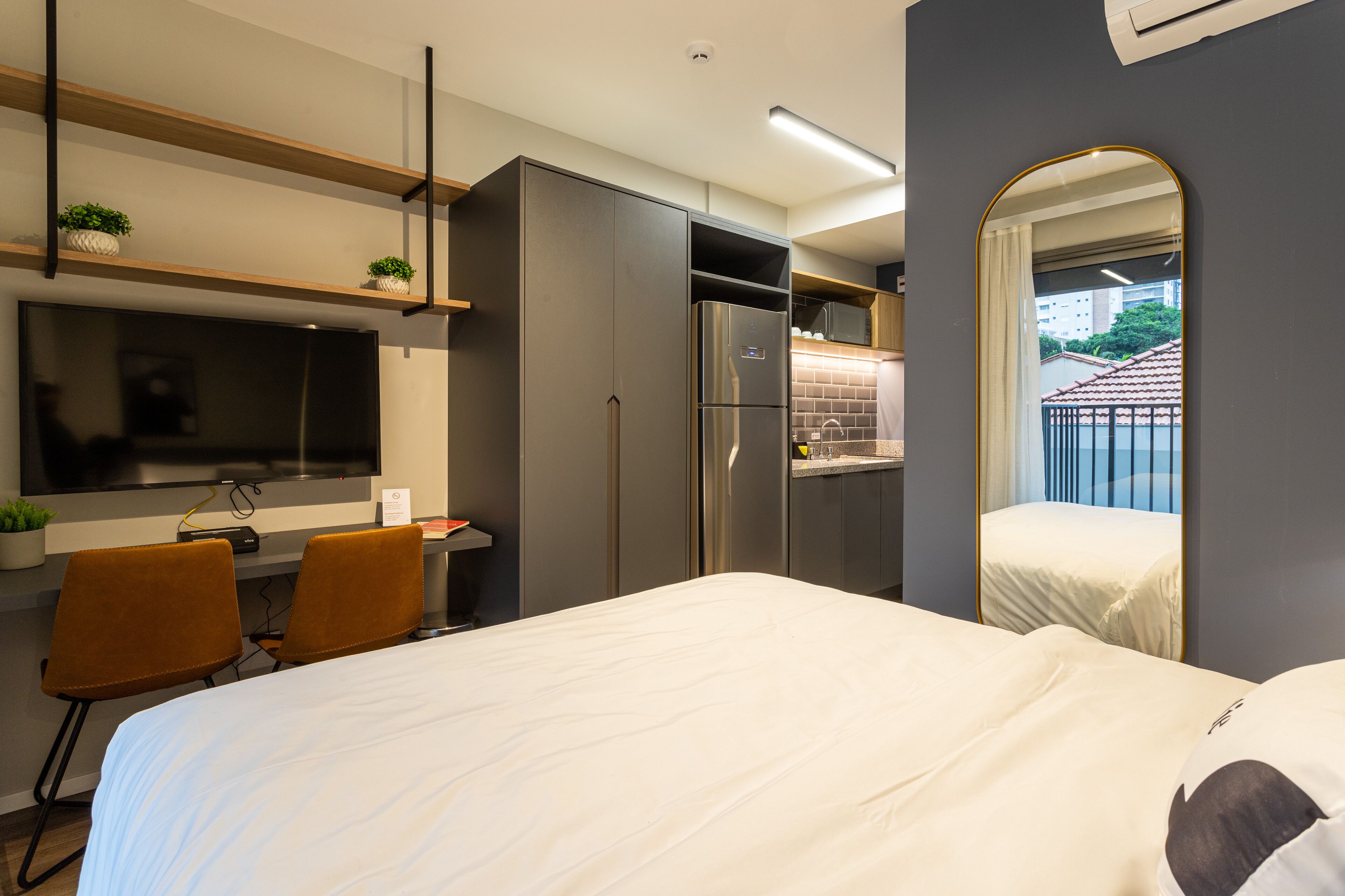 Una habitación de hotel moderna con cama, escritorio con silla, televisor, refrigerador y baño con espejo.
