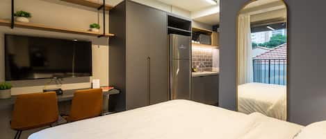 Una habitación de hotel moderna con cama, escritorio con silla, televisor, refrigerador y baño con espejo.