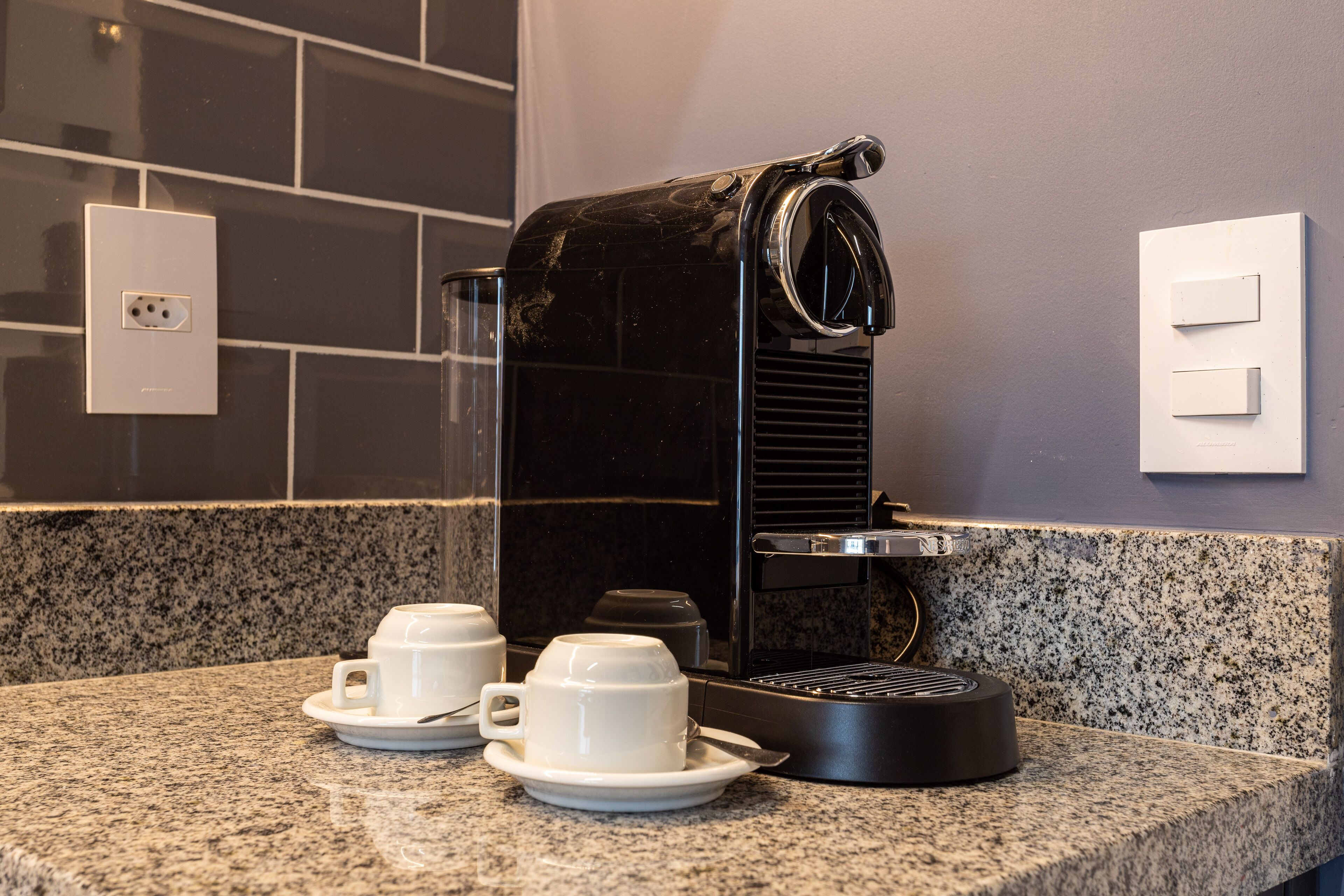 A coffee machine on a granite countertop with two white cups and saucers, a power outlet, and a light switch on the wall.