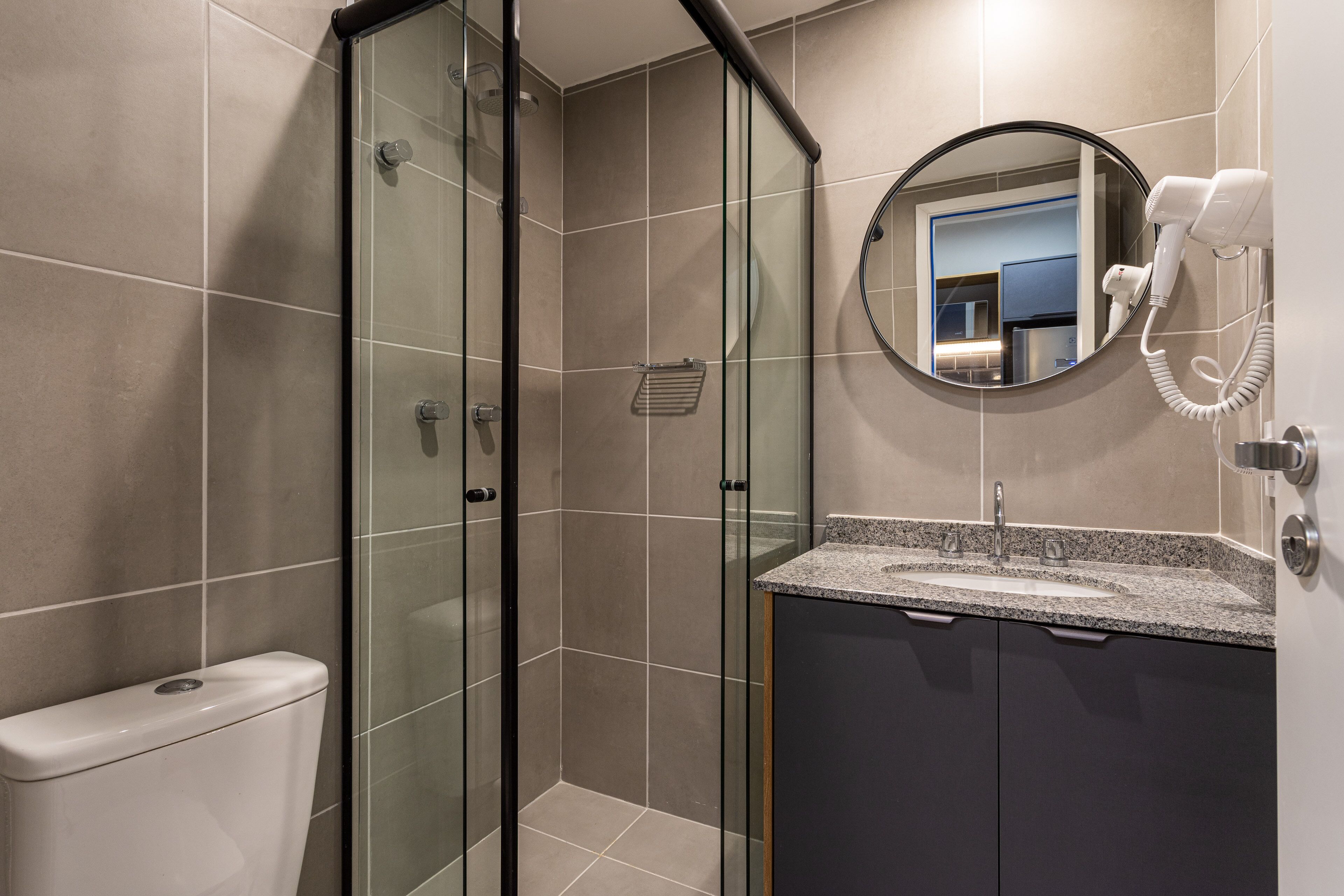 Une salle de bain moderne dotée d’une douche à parois de verre, de toilettes, d’un lavabo avec un plan de travail en granit et d’un sèche-cheveux fixé au mur.