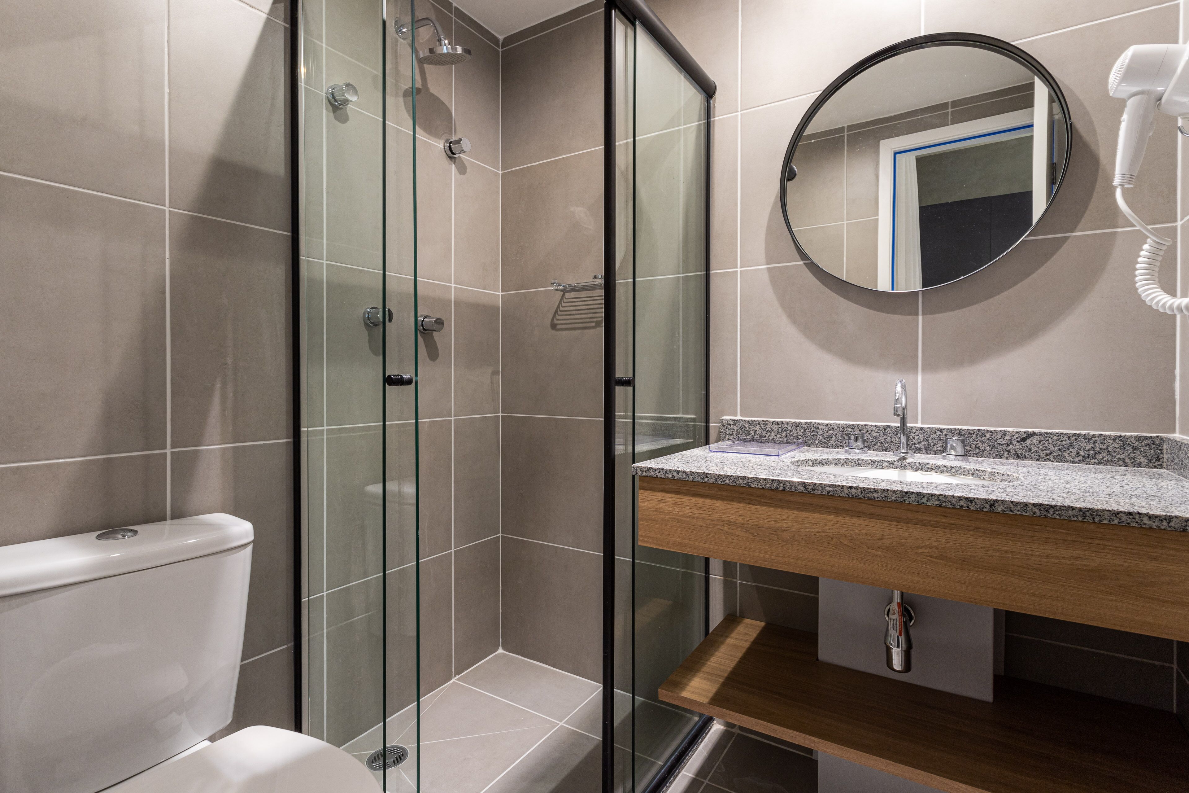 Et moderne badeværelse med et glasafskærmet brusebad, toilet, vask med træbordplade og et rundt spejl på væggen.