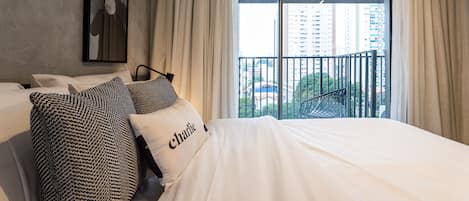 A hotel room with a large bed, a framed picture on the wall, and a balcony with a view of buildings and greenery.