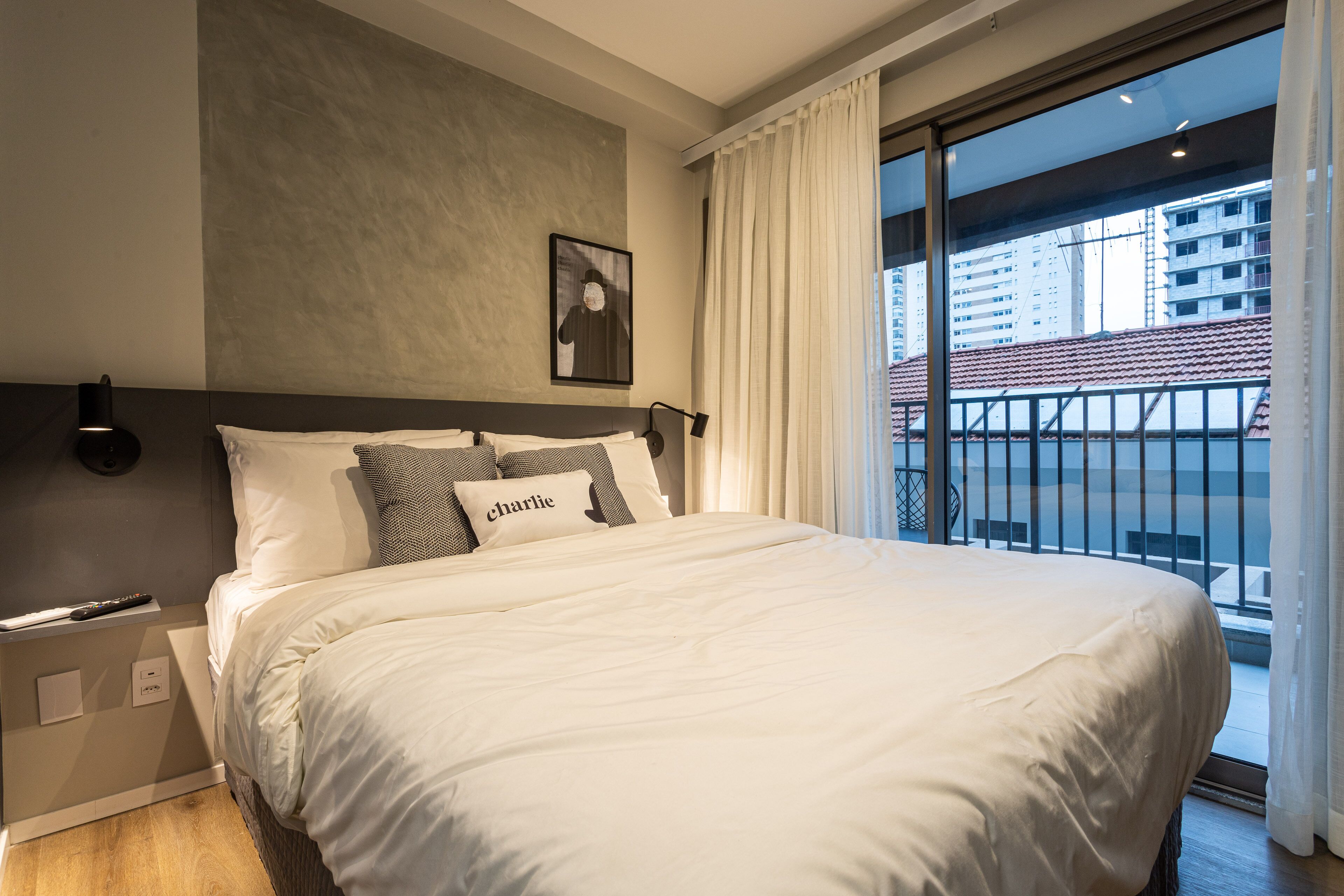 A neatly made bed with white bedding and a pillow labeled "charlie" in a modern hotel room with a large window and a view of buildings.