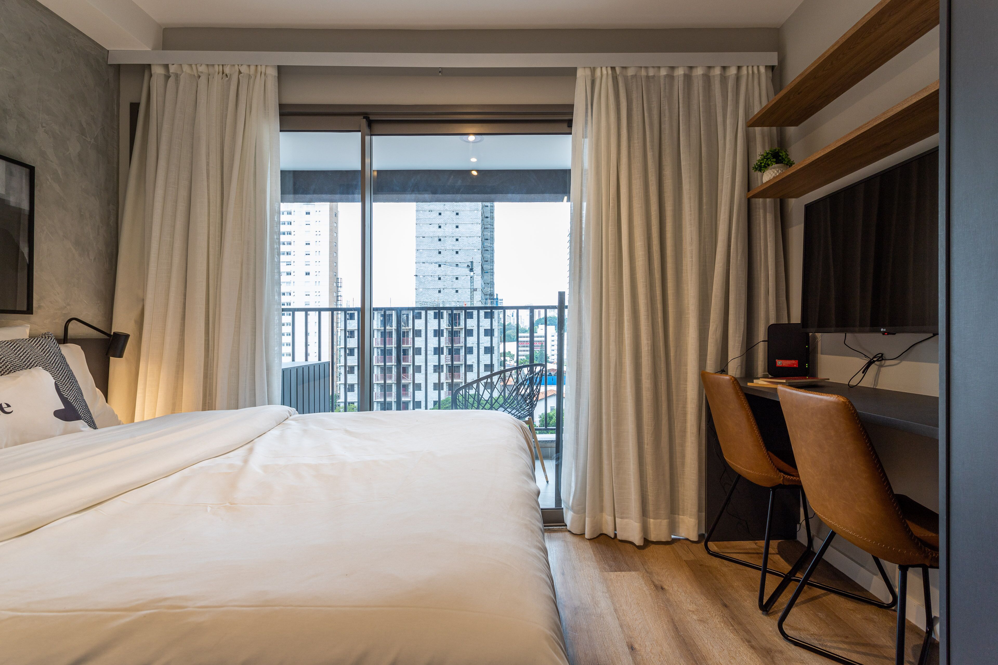 Una habitación de hotel moderna con una cama grande, un escritorio con dos sillas, un televisor y un balcón con cortinas.