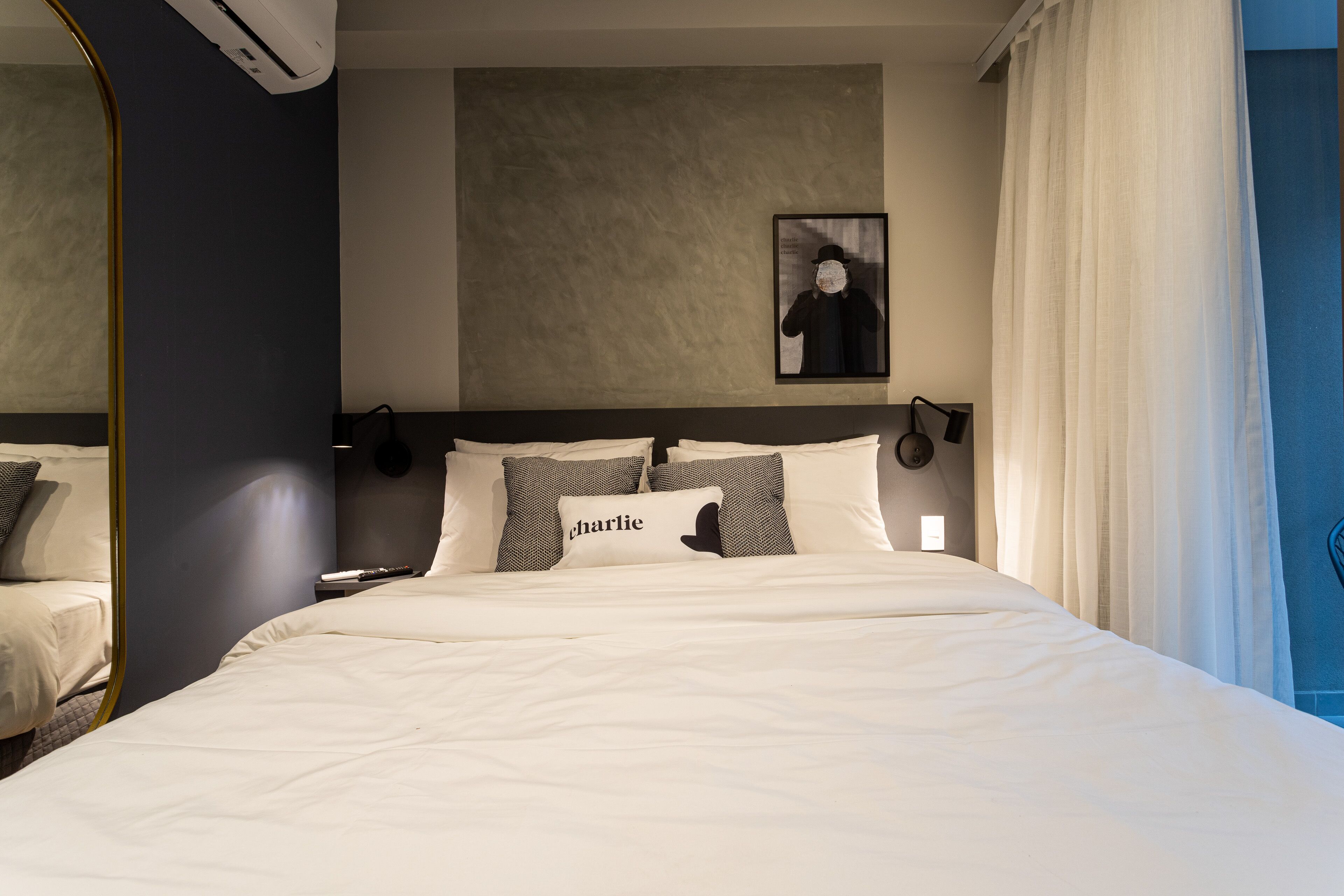 A hotel room with a large bed, a headboard with wall-mounted lights, a framed picture on the wall, and a mirror on the left.