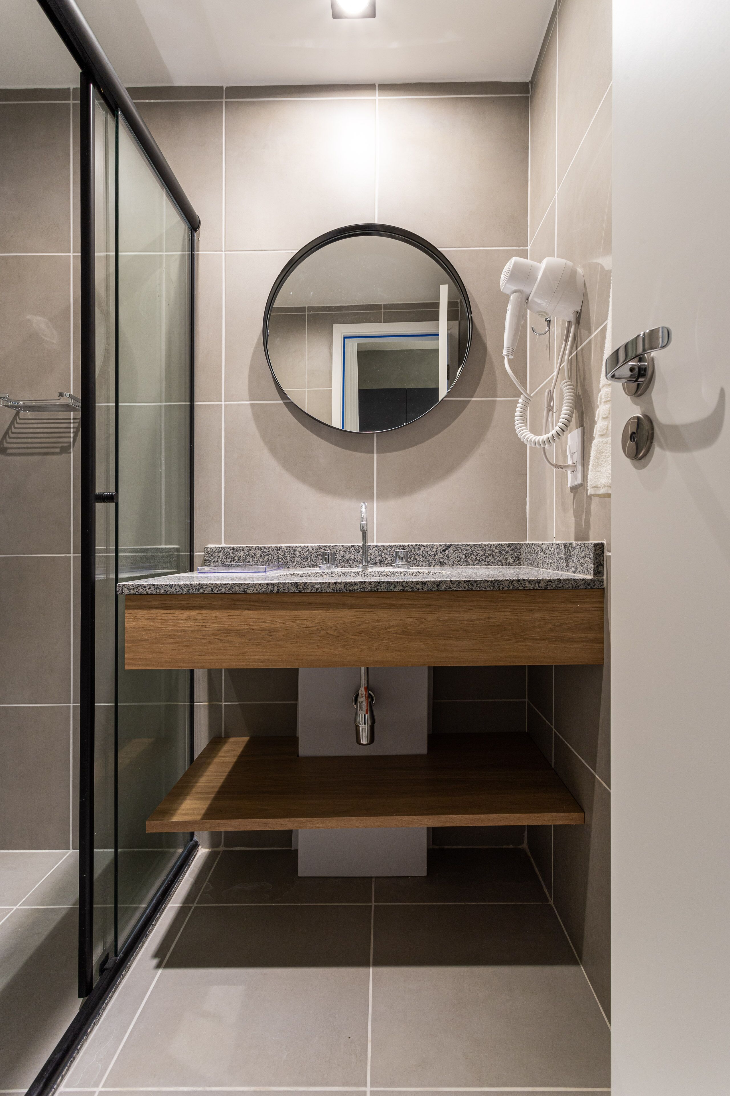 A modern bathroom with a glass shower enclosure, a wooden vanity, a round mirror, and a hairdryer mounted on the wall.