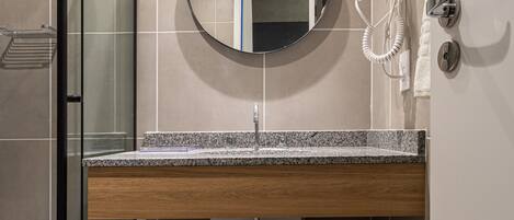 A modern bathroom with a glass shower enclosure, a wooden vanity, a round mirror, and a hairdryer mounted on the wall.