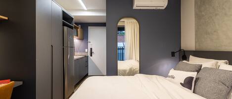 A modern hotel room with a large bed, a built-in wardrobe, a kitchenette, and a view through an arched doorway.