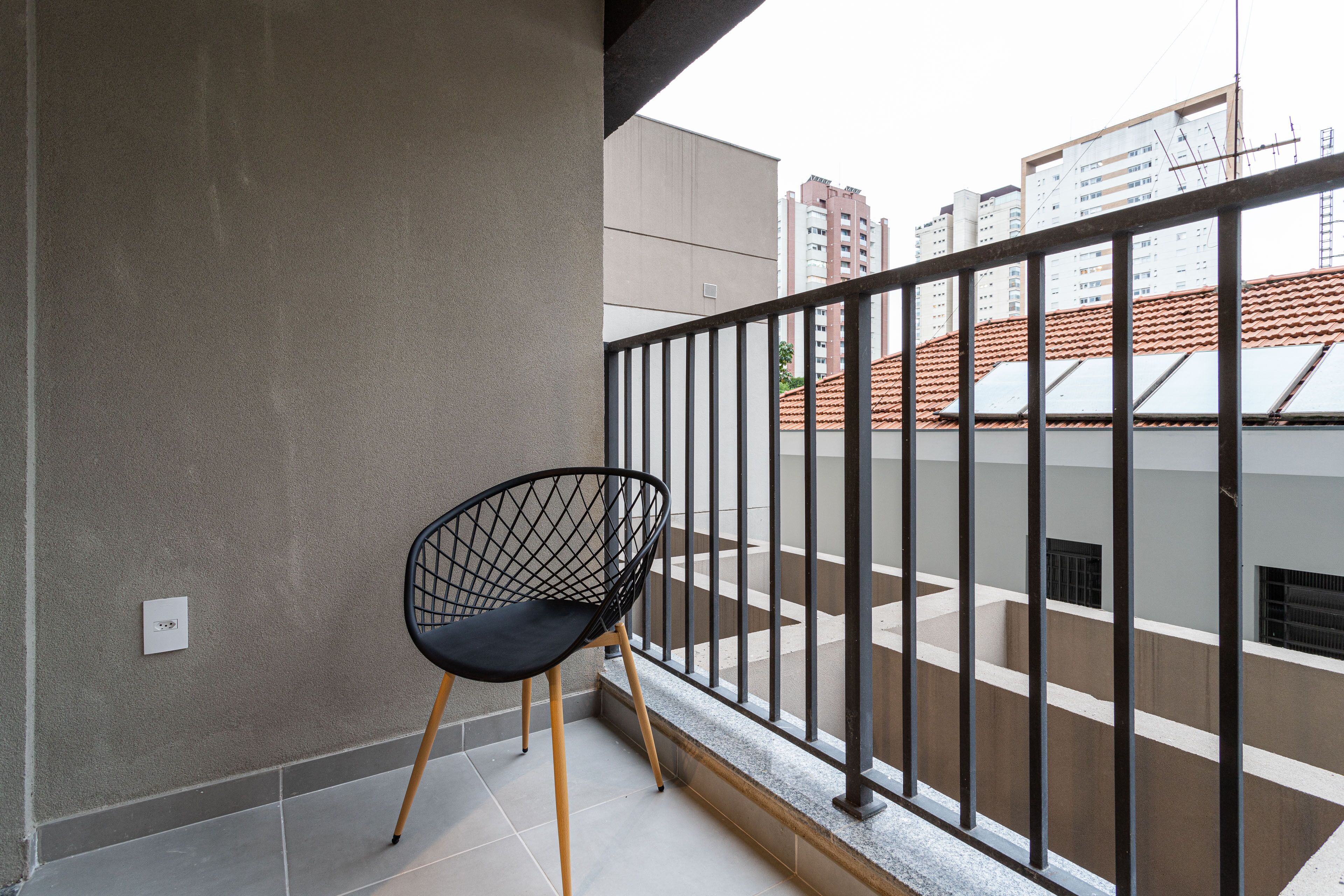 A balcony with a black wireframe chair, a tiled floor, and a view of a building with a red-tiled roof.